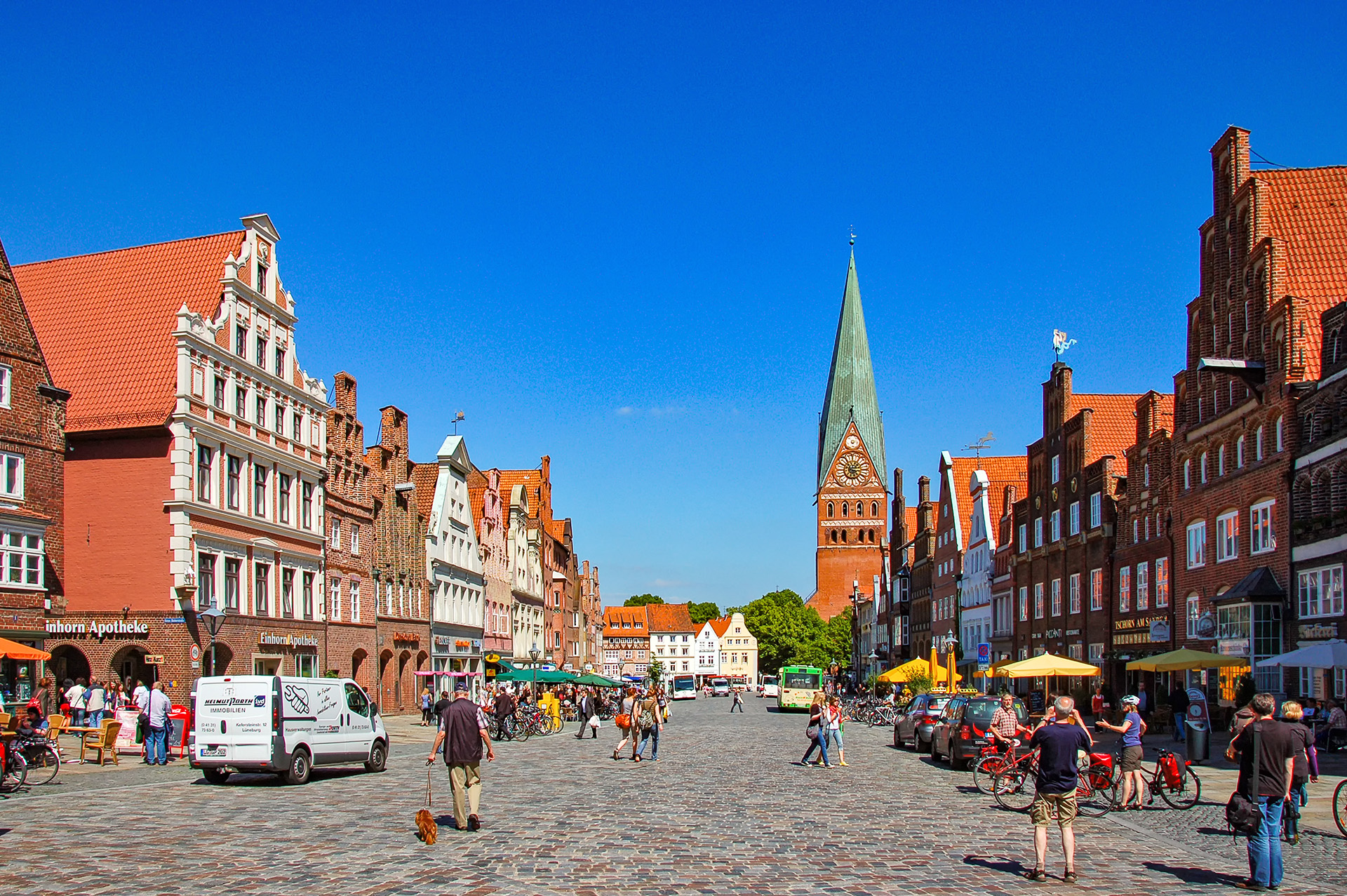 Der Platz Am Sande ist einer der ältesten Plätze in Lüneburg. Hier sind viele Fachwerkhäuser mit spitzen Giebeln und der imposante Kirchturm der St. Johanniskirche zu finden. Die Außengastronomie zieht viele Besucher:innen auf den Platz an.